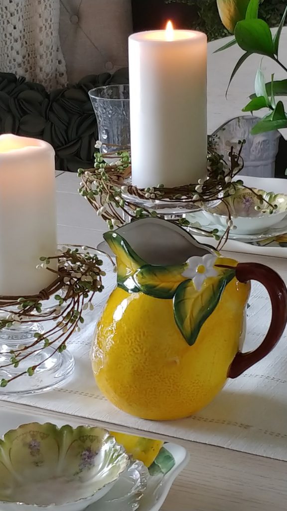 A bowl of fruit on a table, with Yellow pitcher and candles