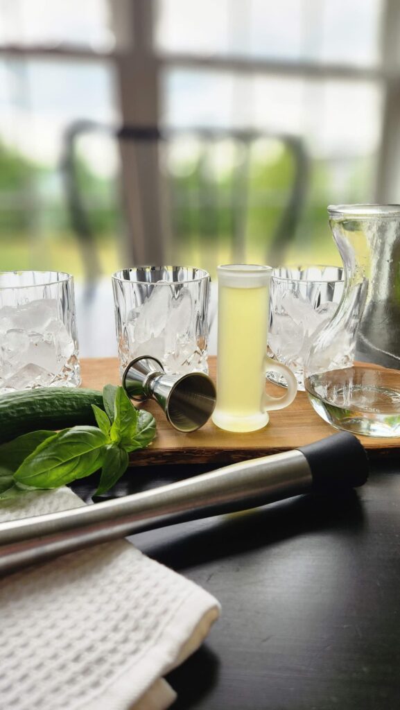 muddler with three glasses, basil and shot glass of limoncello to make a cocktail on a table