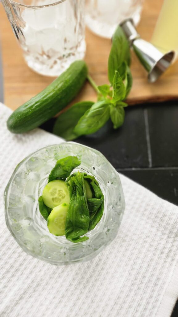 overhead photo of glass  with basil and cucumber in the bottom after being muddled