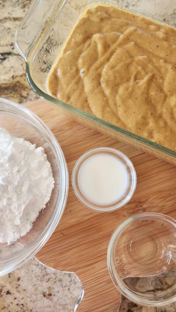 pumpkin bread batter in load pan with powdered sugar and milk in small glass bowls next to loaf pan