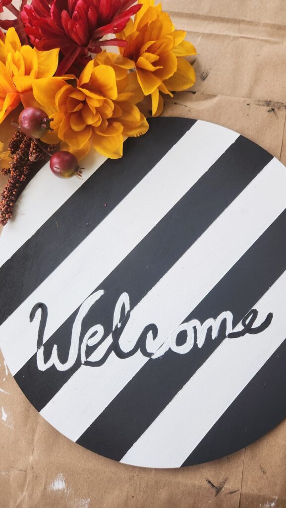 round wooden door hanger painted with stripes and handwritten welcome painted on it