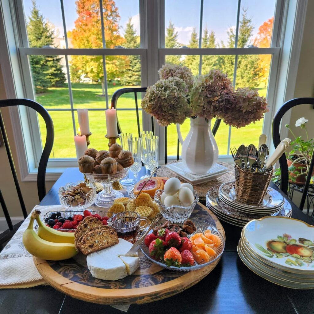 view of kitchen table in front of window with a breakfast charcuterie board on top filled with breakfast foods