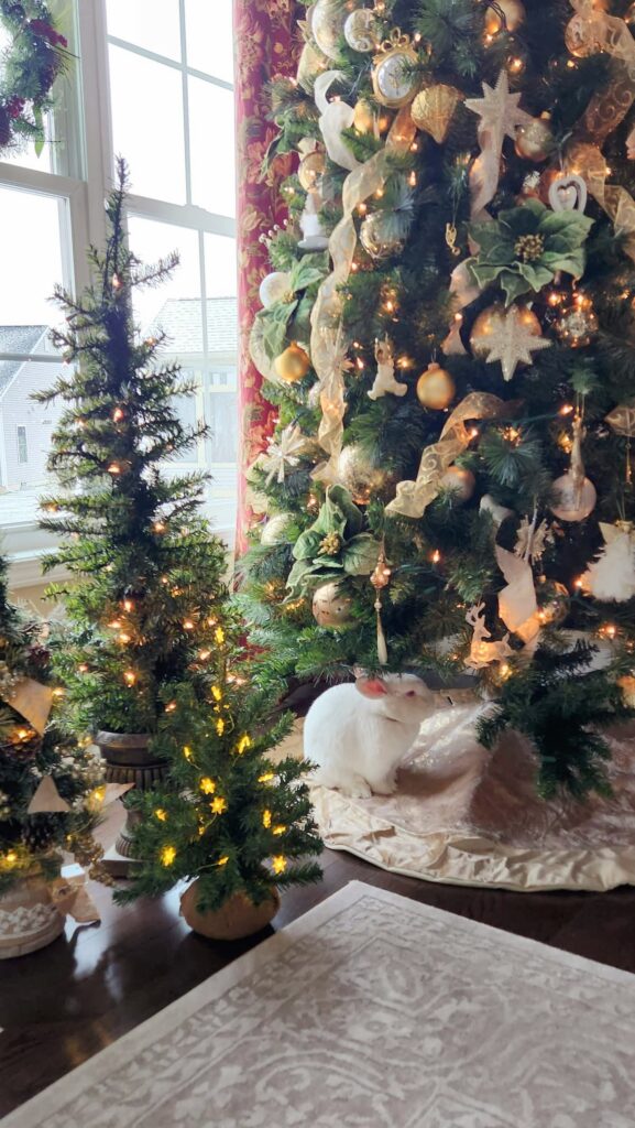 Christmas tree with gold and ivory ornaments in front of window with white bunny rabbit sitting underneath the tree
