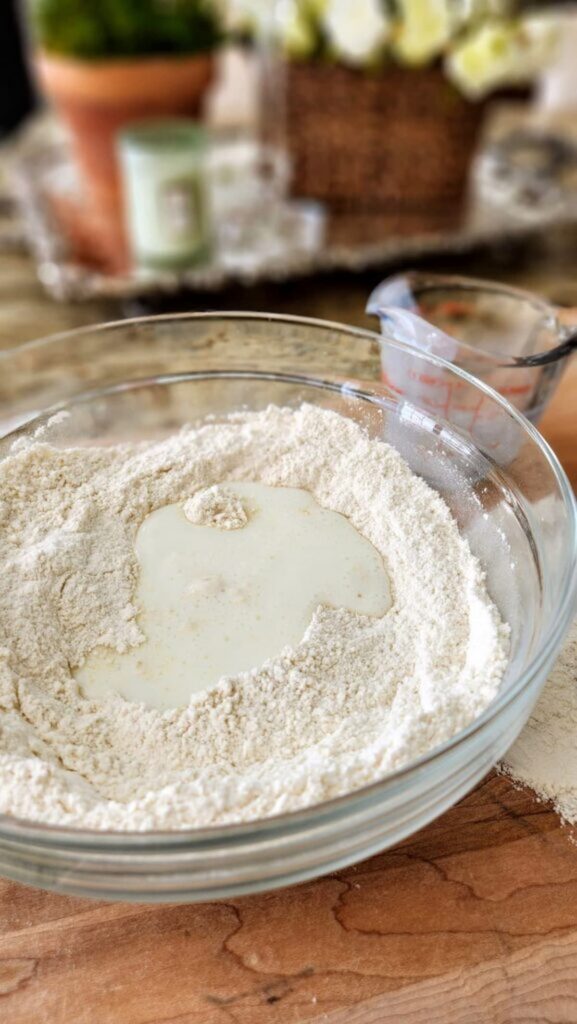 glass mixing bowl with a cup of buttermilk in center of flour that is in the bowl