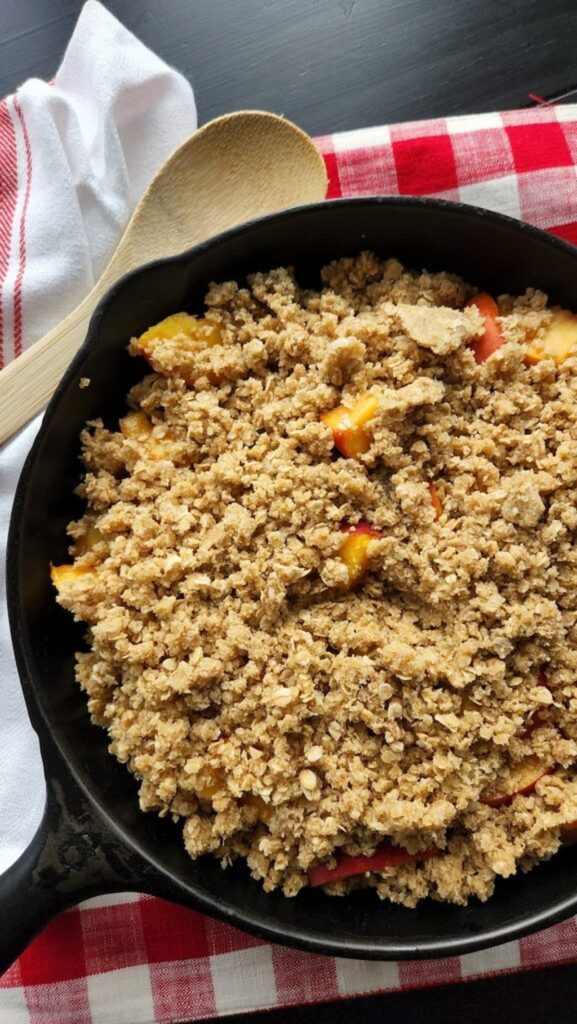 overhead view of cast iron skillet with peach crisp in it and wooden spoon on side of pan