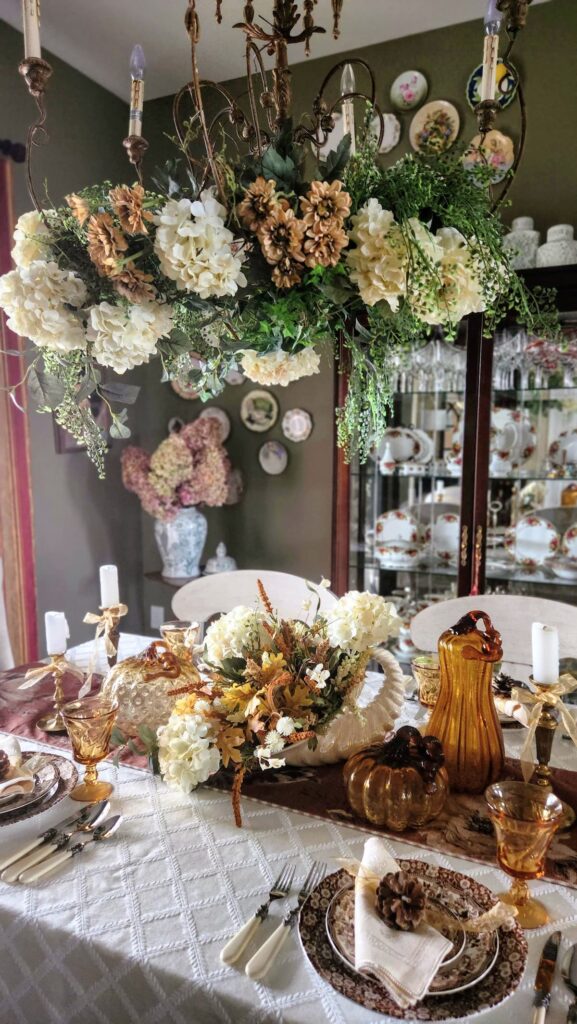 view of dining room table set for thanksgiving with neutral creams, browns and charndelier with flowers and greeenery hanging from it