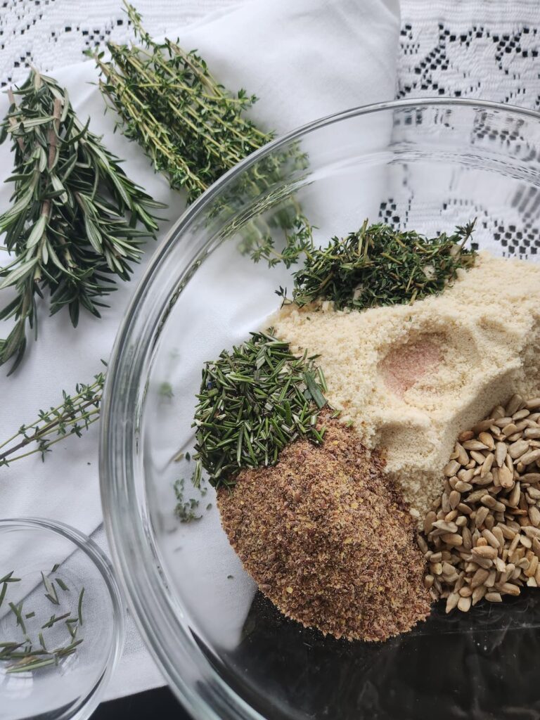 overhead of glass bowl with sunflower seeds, flax seeds, almond flour and fresh herbs for gluten free crispbread recipe