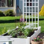 White raised bed spring garden with trellis iron obelisk foxglove and pink dahlia container beside Adirondack chair