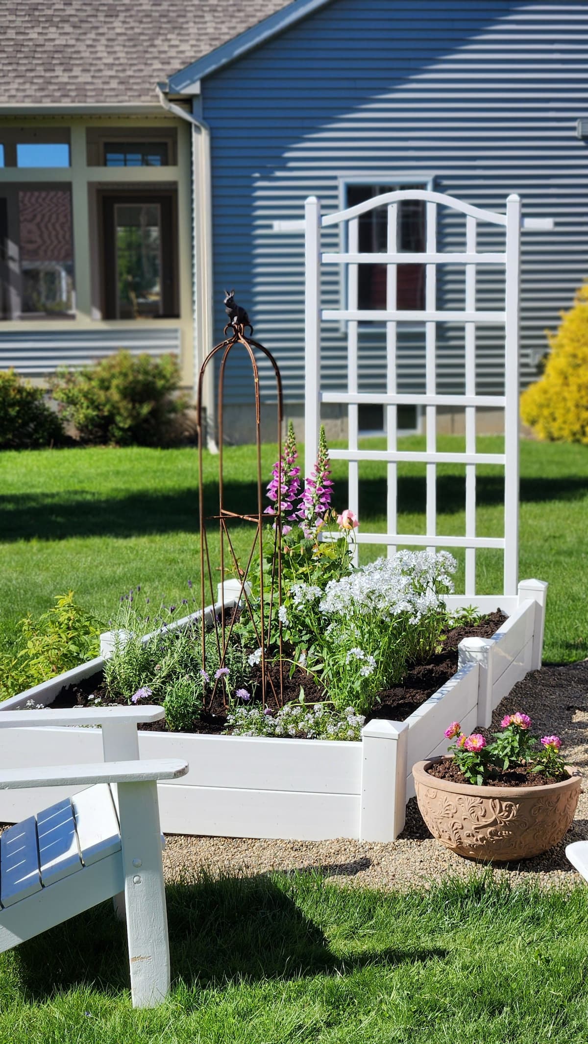 White raised bed spring garden with trellis iron obelisk foxglove and pink dahlia container beside Adirondack chair