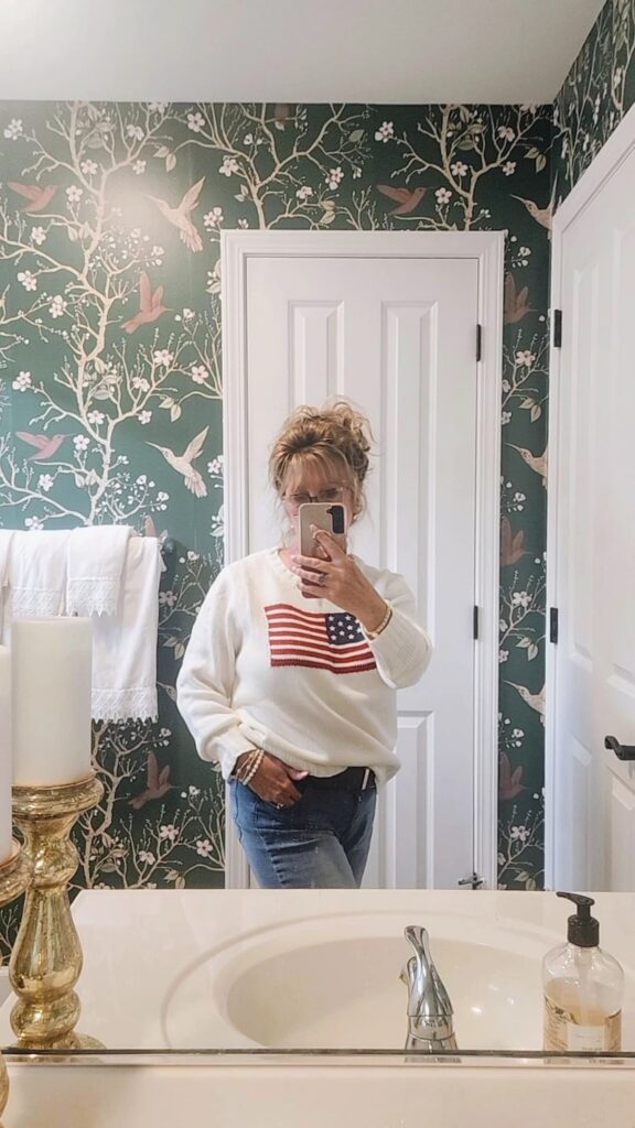 Mirror selfie wearing a cream American flag sweater with jeans in a bathroom with dark green hummingbird wallpaper