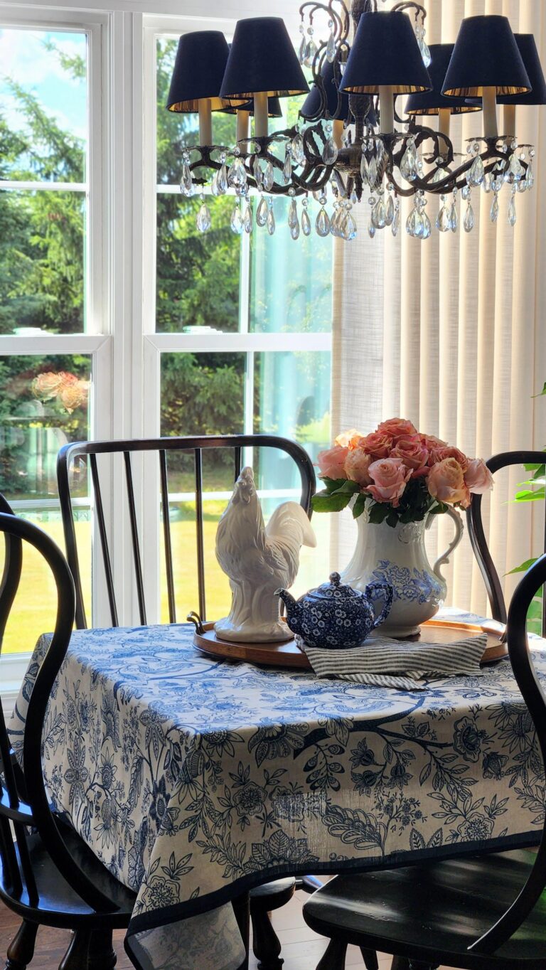 French country dining room with blue and white toile tablecloth, peach roses in an ironstone pitcher, white ceramic rooster, and antique bronze crystal chandelier — simple spring home decorating ideas