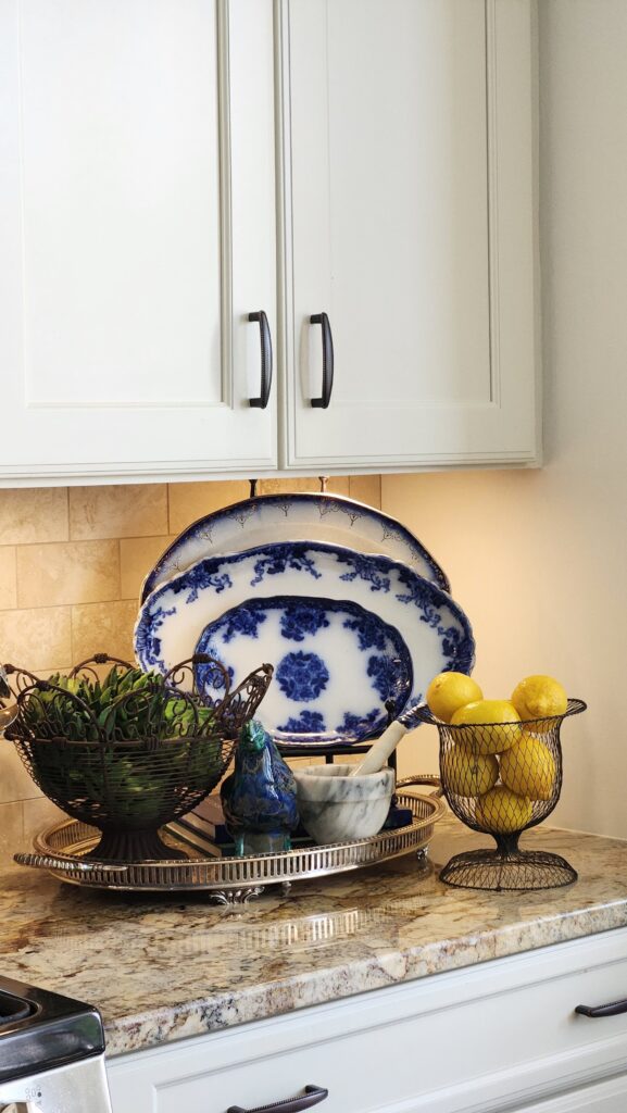  French country kitchen counter vignette with stacked antique flow blue platters, wire baskets holding artichokes and lemons, a marble mortar and pestle, and a ceramic bird on a silver tray