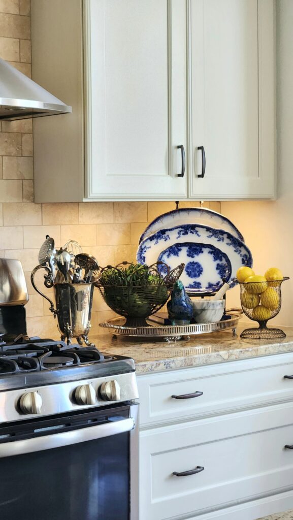 French country kitchen decorated with antique flow blue platters, wire fruit baskets, silver utensil holder, and lemon-filled wire compote displayed on a granite countertop beside a gas stove