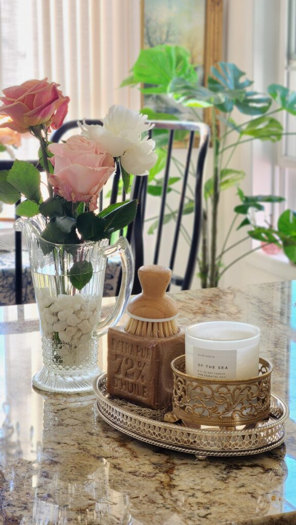 Peach and blush roses with a white peony arranged in a vintage crystal pitcher on a granite kitchen counter beside a French Savon de Marseille soap block and bamboo brush