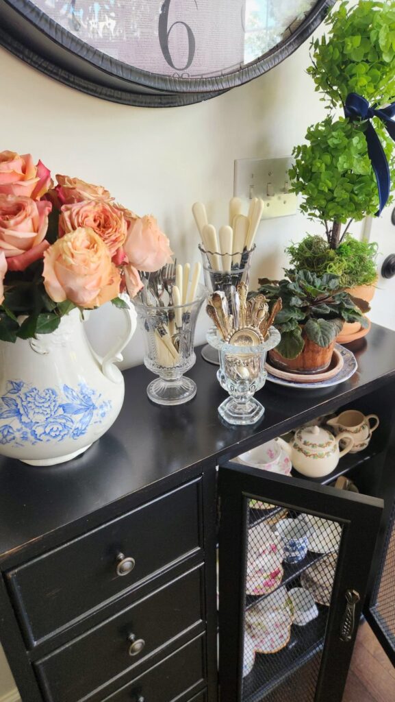  Simple Spring Home Tips Black sideboard vignette with peach garden roses in a blue and white ironstone pitcher, vintage crystal compotes holding ivory-handled and gold flatware, potted plants, and antique tea sets displayed inside a mesh cabinet below