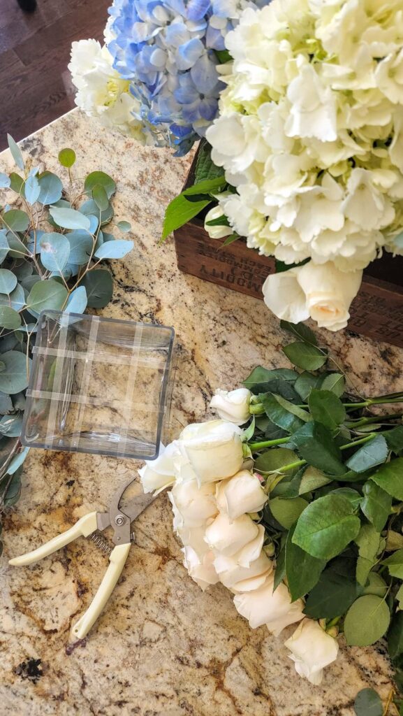 Flat lay of Trader Joes flowers being arranged with white roses hydrangea eucalyptus scissors and clear vase on granite counter