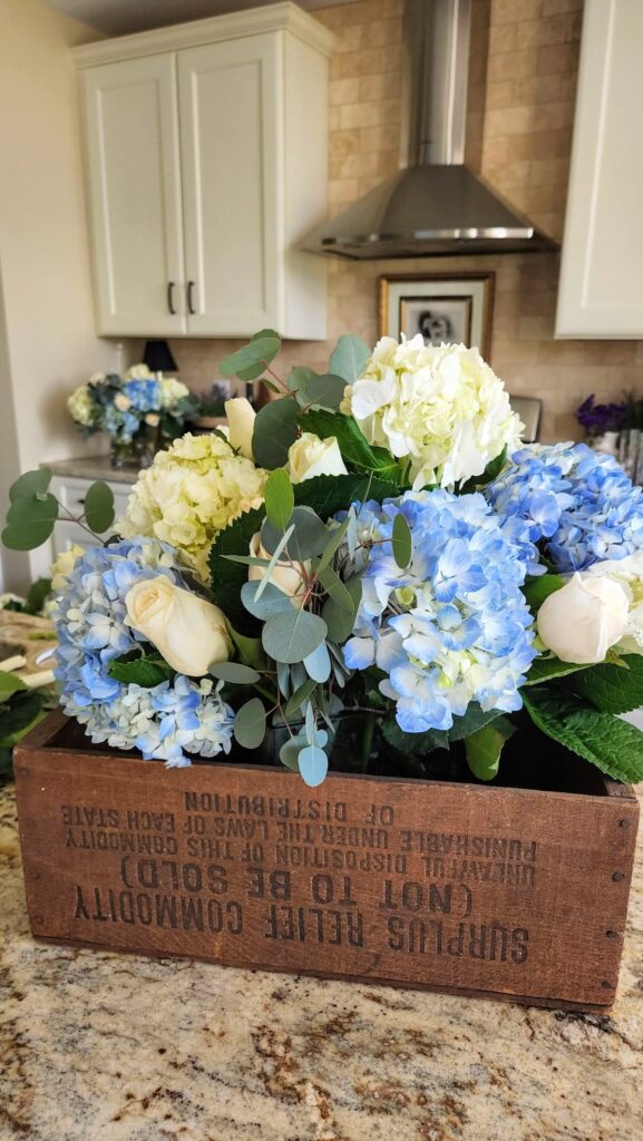 Cream roses laying on granite counter with blue and white hydrangea arrangement in vintage wooden crate