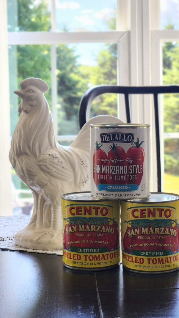White ceramic rooster figurine on a lace doily beside Cento and DeLallo San Marzano tomato cans on a dark wood table